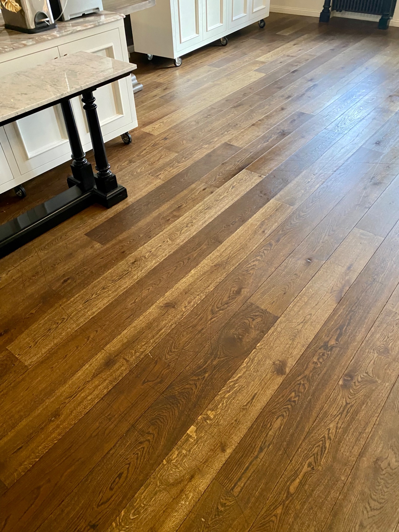 Ukośne ujęcie brązowej podłogi z desek w kuchni, widoczne białe szafki z marmurowym blatem i czarny stół na ozdobnych nogach.