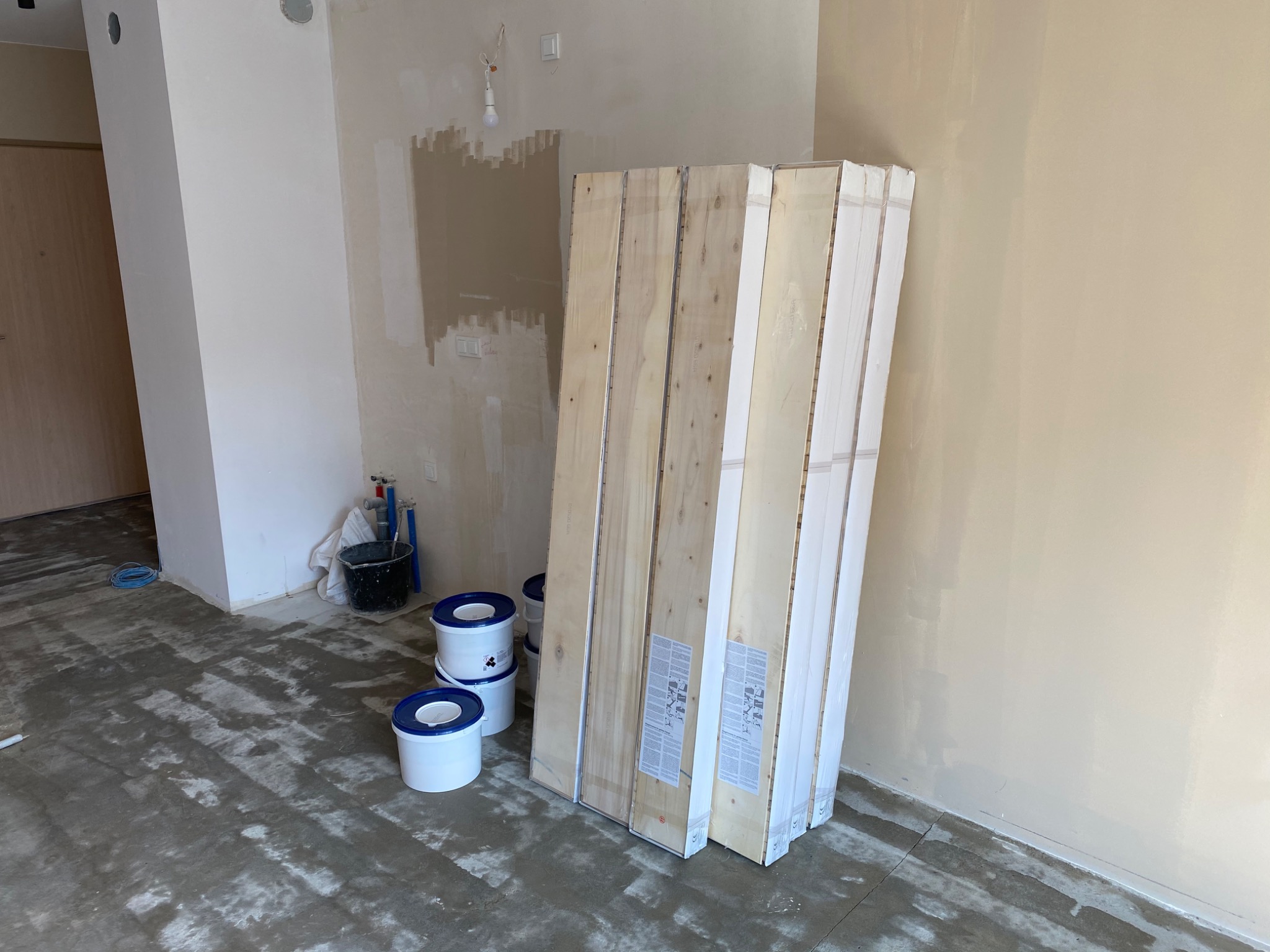 Przygotowanie do montażu: Paczki paneli podłogowych o jasnym drewnie, stojące w surowym wnętrzu, obok wiader z klejem i masą szpachlową na wylewce samopoziomującej.