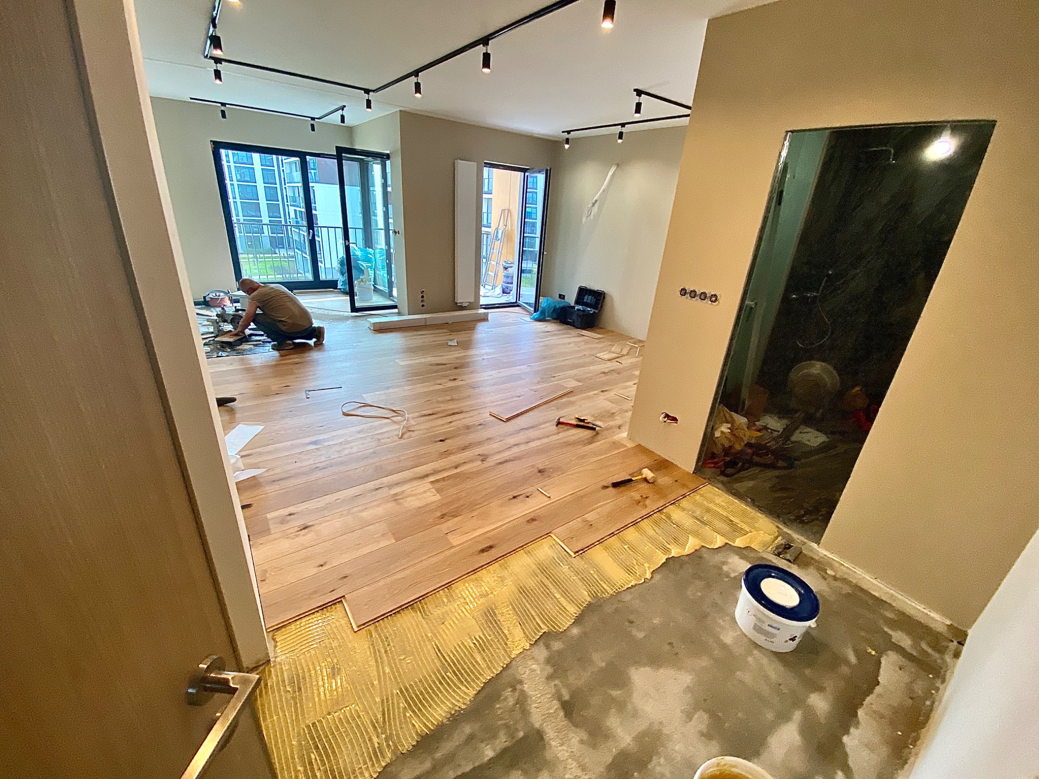 Proces montażu jasnych desek podłogowych w przestronnym apartamencie z widokiem na miasto, widoczny pracownik w trakcie docinania elementu przy użyciu narzędzi.