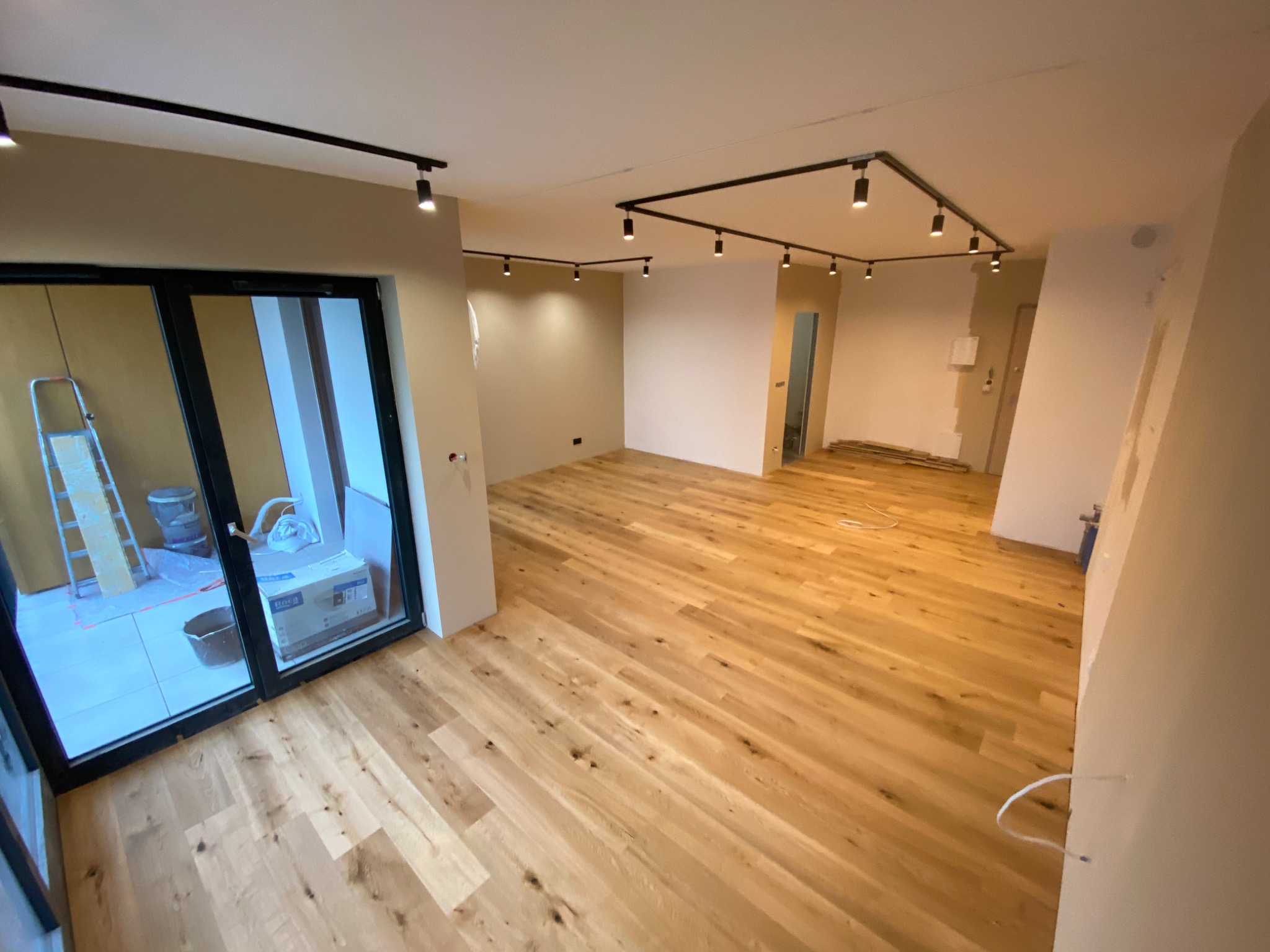 Jasny, świeżo ułożony parkiet w przestronnym salonie z oświetleniem szynowym na suficie. Widoczne przygotowania do dalszych prac wykończeniowych, w tle drabina i materiały budowlane za...