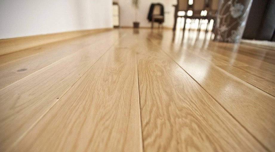 Jasne, lakierowane deski parkietowe ułożone wzdłużnie, z widocznym usłojeniem drewna i odbiciami światła, prowadzące w głąb jasnego pomieszczenia z elementami wyposażenia w tle.