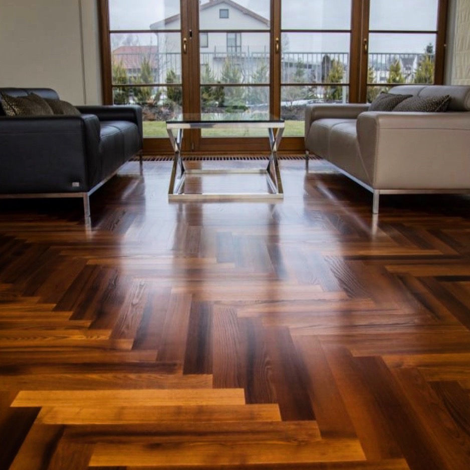 Parkiet ułożony we wzór jodełki francuskiej, wykończony lakierem o wysokim połysku, odbijający światło z dużego okna tarasowego, w tle dwa nowoczesne fotele i szklany stolik kawowy.