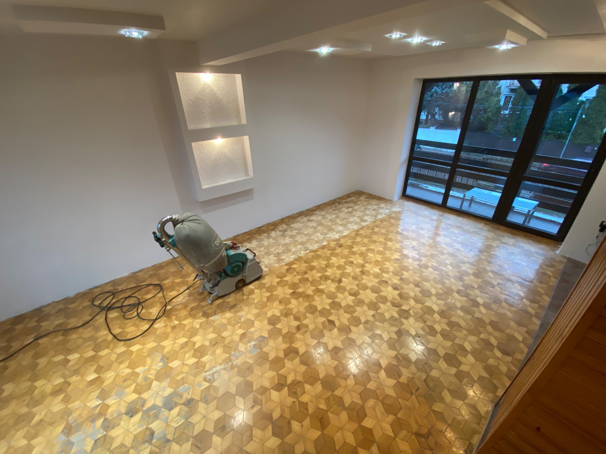 Parkiet ułożony we wzór 3D, częściowo odnowiony za pomocą szlifierki podłogowej, widoczne różnice w kolorze i połysku między obszarami obrobionymi i nieobrobionymi.