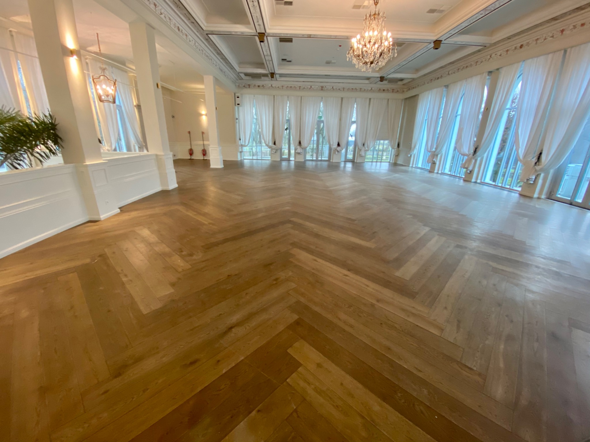 Jasna, przestronna sala z parkietem ułożonym we wzór jodełki francuskiej, wysokie okna z białymi zasłonami, kryształowy żyrandol i białe kolumny.