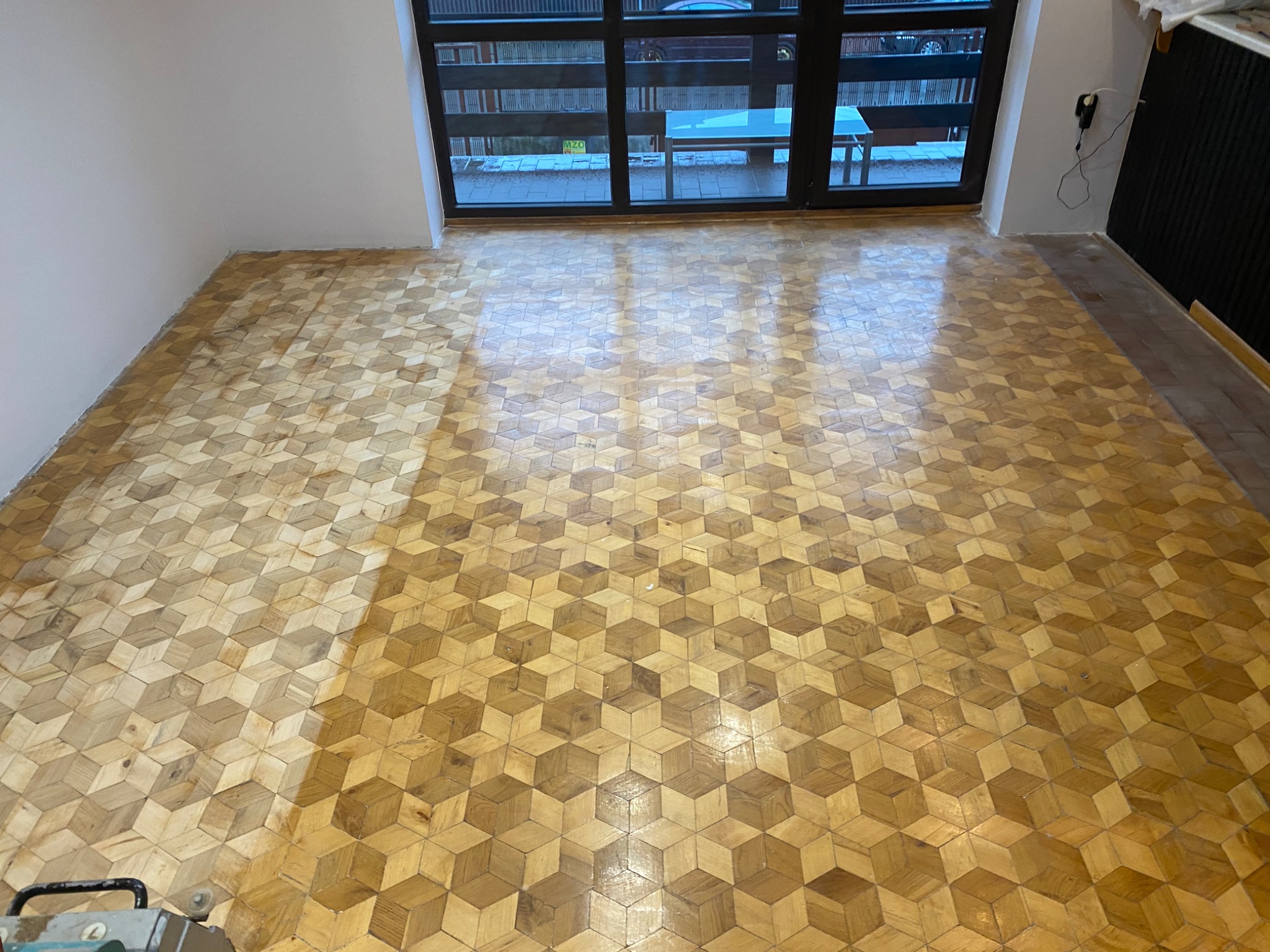 Parkiet ułożony we wzór kostki 3D, częściowo odnowiony, z widocznym urządzeniem do renowacji w rogu pomieszczenia, duże okno w tle.