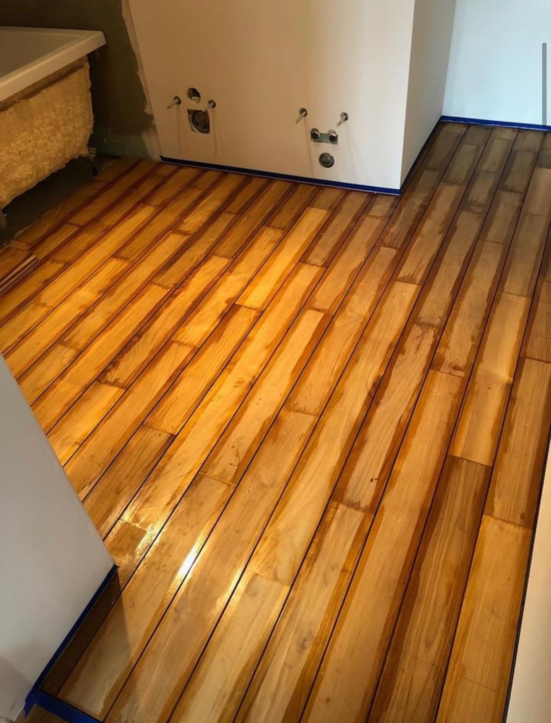 Świeżo lakierowana podłoga drewniana z widocznymi smugami lakieru, zabezpieczona niebieską taśmą malarską przy ścianach w trakcie renowacji łazienki.