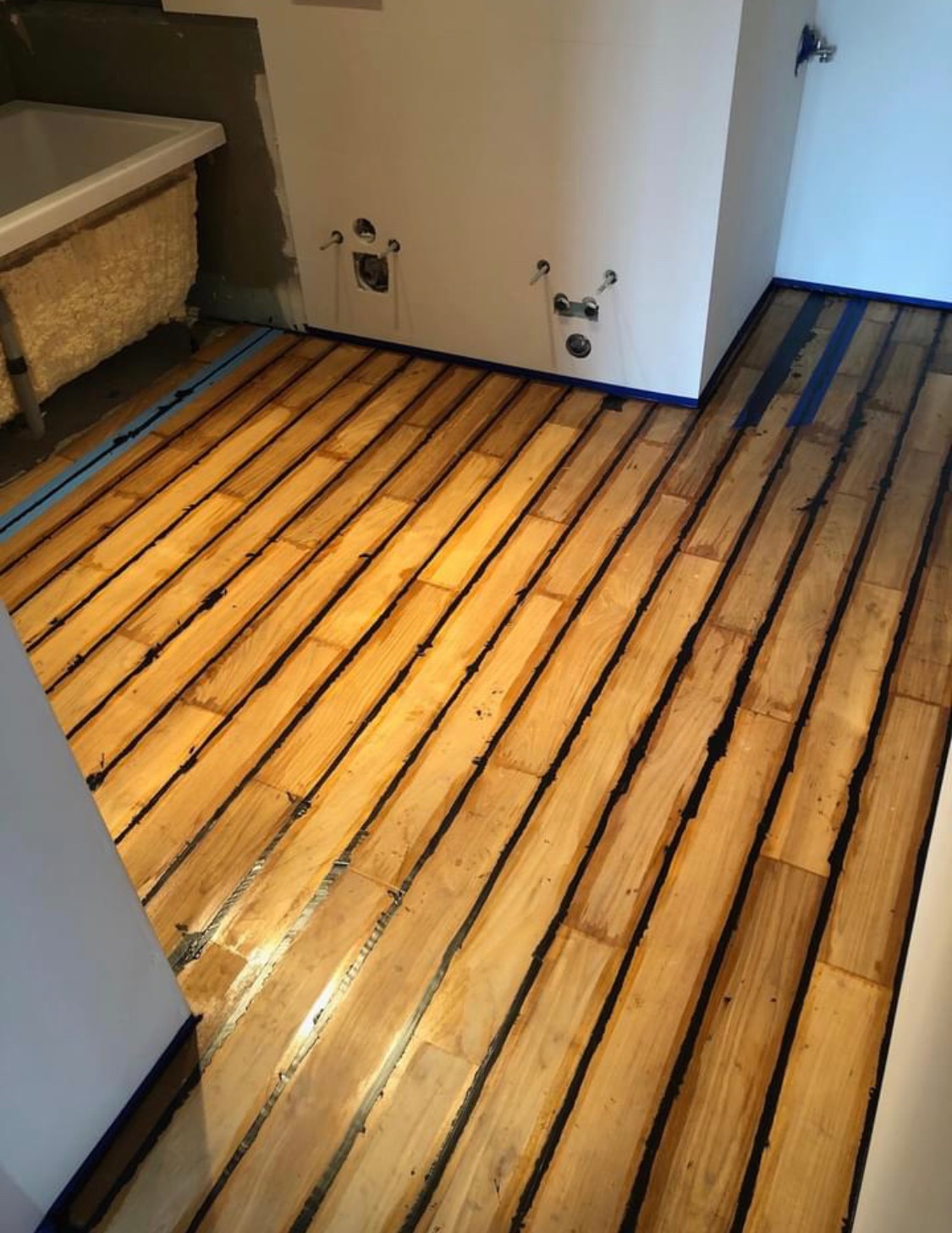 Przygotowane do lakierowania deski podłogowe z widoczną taśmą malarską wzdłuż ściany, fragment wanny z pianką montażową w tle.