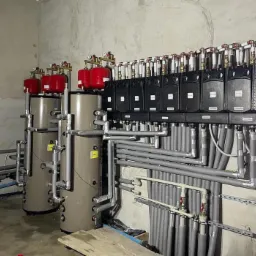 Instalacja centralnego ogrzewania z szarymi zbiornikami akumulacyjnymi, czerwonymi naczyniami przeponowymi i czarnymi rozdzielaczami, rury izolowane szarą otuliną, zamontowana w surowym pomieszczeniu.