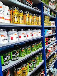 Półki sklepowe wypełnione różnorodnymi suplementami diety, w przeważającej mierze kreatyną, w opakowaniach o intensywnych kolorach i różnych rozmiarach, ustawione równo na niebieskich półkach.