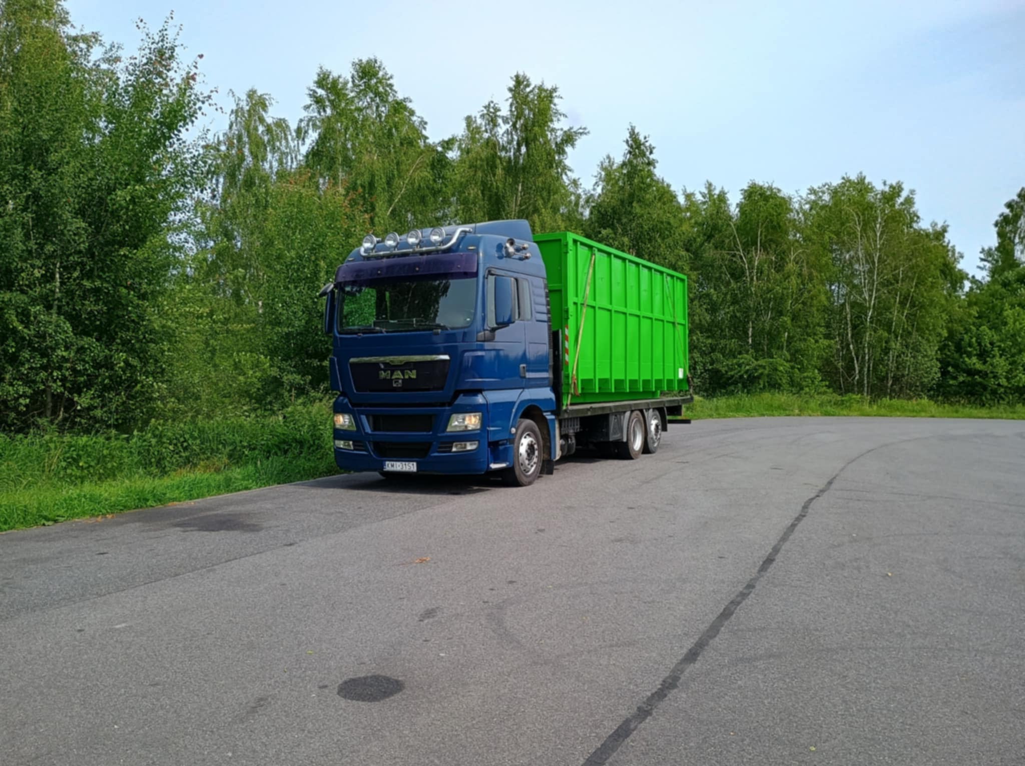 Niebieska ciężarówka MAN z zielonym kontenerem na szosie, otoczona zielenią drzew. Widok z boku, lekko z dołu, z widocznym numerem rejestracyjnym pojazdu.