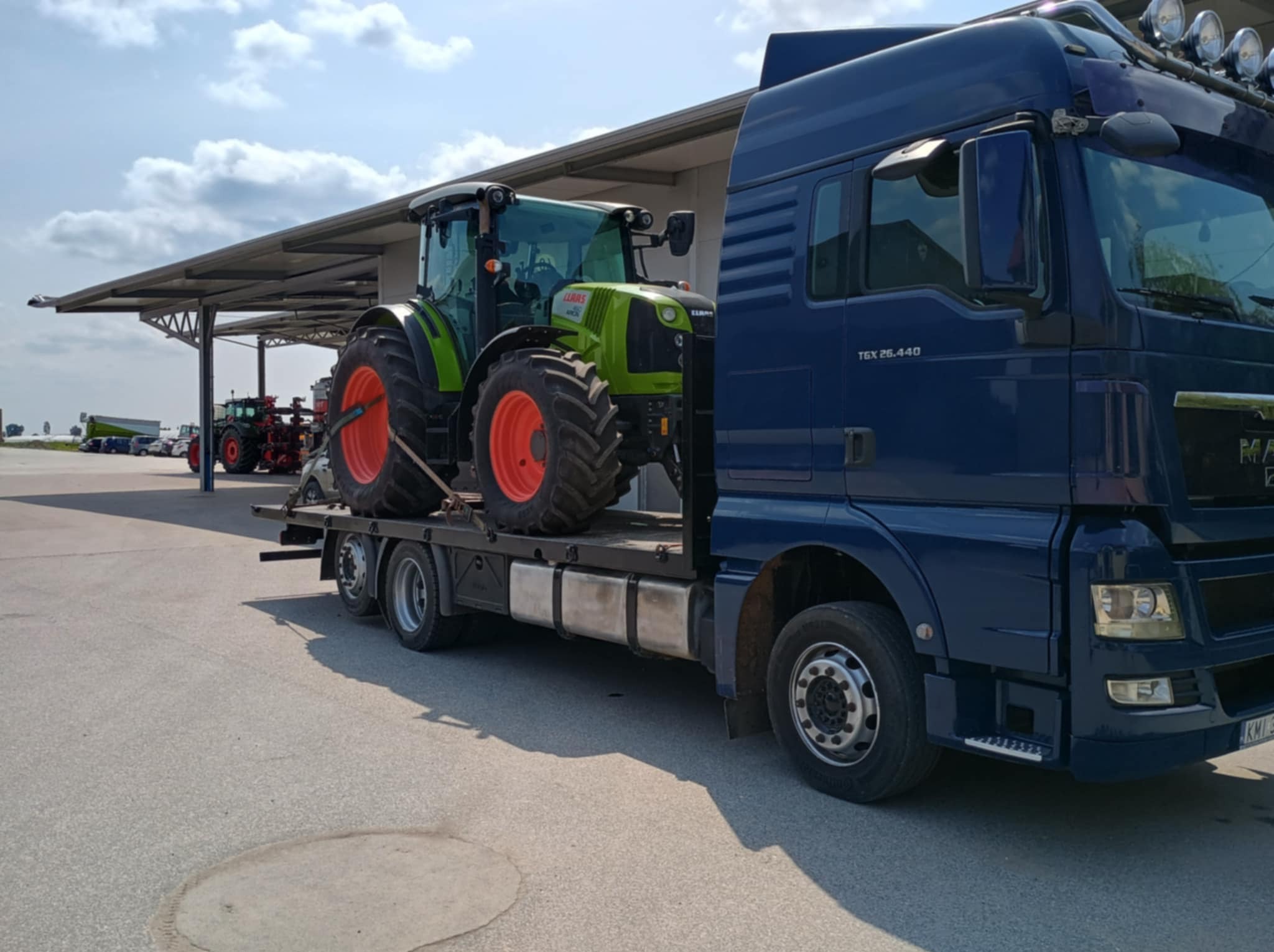 Niebieska laweta MAN z zielonym traktorem Claas na platformie. Widoczne inne traktory w tle, pod zadaszeniem. Słoneczny dzień.