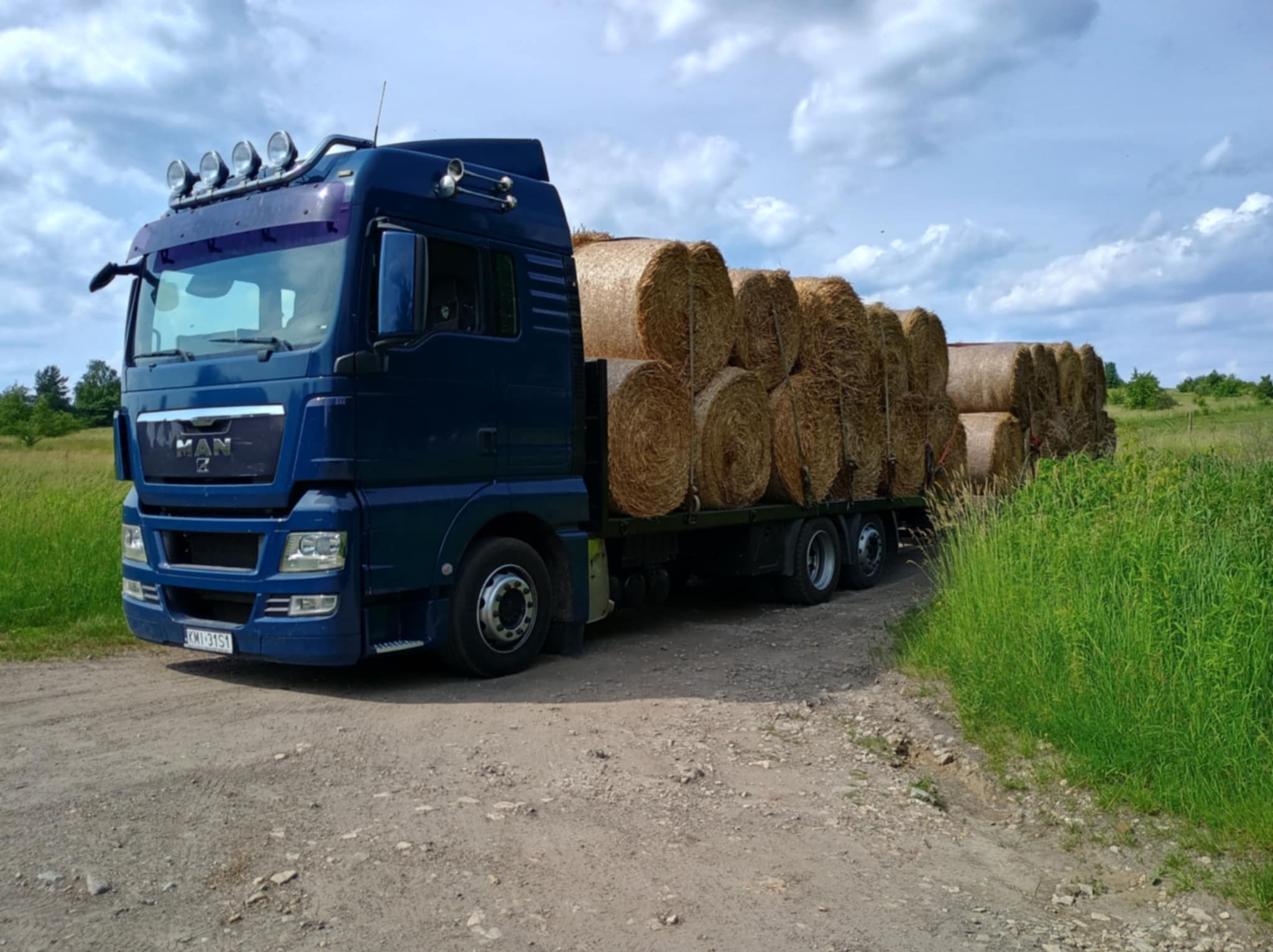 Niebieska ciężarówka MAN z ładunkiem okrągłych bel siana na przyczepie, widziana z boku na polnej drodze, w tle łąka i błękitne niebo z chmurami.