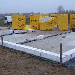 Fundament domu jednorodzinnego w trakcie budowy, z widocznymi bloczkami Ytong, izolacją poziomą i zapasem materiałów budowlanych w tle, na zewnątrz w pochmurny dzień.