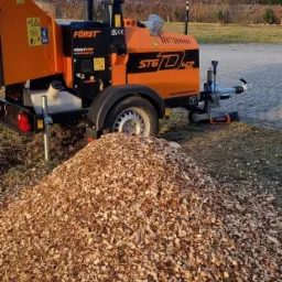 Pomarańczowa rębarka do gałęzi marki Först ST6D42 z czarnym kominem i przyczepą stoi obok dużej pryzmy zrębków na trawiastym terenie.