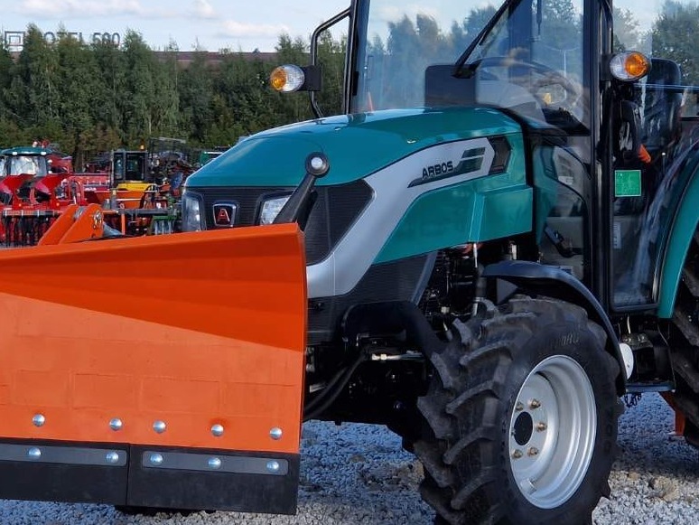 Zielono-srebrny ciągnik Arbos z pomarańczowym pługiem do śniegu, widok z przodu, opony z wyraźnym bieżnikiem, w tle inne maszyny rolnicze.