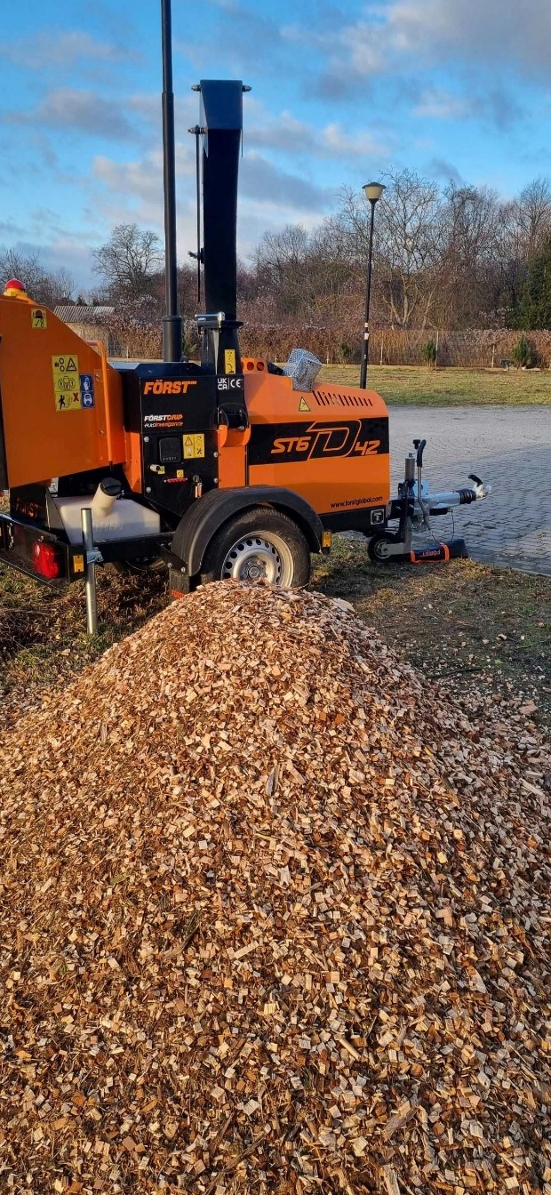 Pomarańczowa rębarka do gałęzi marki Först ST6D42 z czarnym kominem i przyczepą stoi obok dużej pryzmy zrębków na trawiastym terenie.