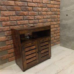 And Bud - Ciemnobrązowa szafka pod umywalkę z lamelowymi drzwiczkami, na tle ściany z cegły klinkierowej i szarych płytek, widoczne otwory po montażu.