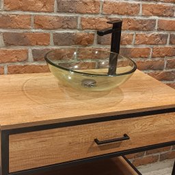 And Bud - Szklana umywalka nablatowa z czarną armaturą na drewnianej szafce z szufladą, tło z cegły klinkierowej z gniazdkiem elektrycznym.