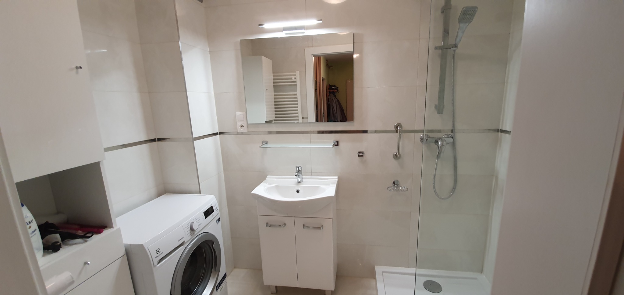 Jasna łazienka z białymi płytkami, kabiną prysznicową ze szklaną ścianką, umywalką z szafką, pralką Electrolux i szafką z półkami