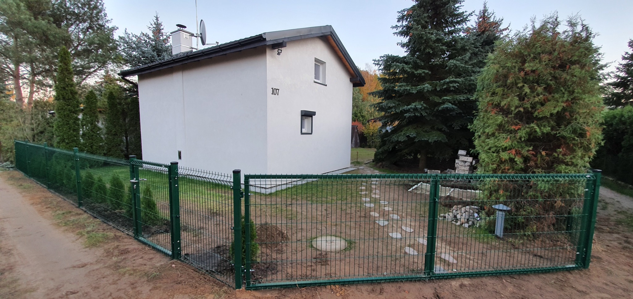 Nowoczesny dom jednorodzinny z zielonym ogrodzeniem panelowym, widoczne ułożone kamienne płyty chodnikowe i trawnik w trakcie zakładania, otoczenie drzew i krzewów.