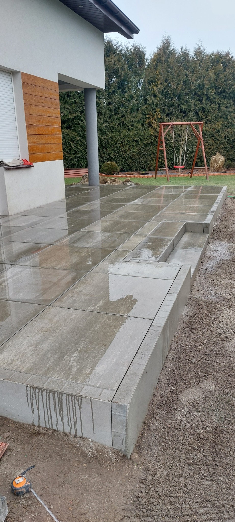 Świeżo ułożona, mokra, betonowa nawierzchnia tarasu z niskimi schodkami, widoczne narzędzia pomiarowe leżące na ziemi, w tle dom z drewnianą elewacją i huśtawka na trawniku.