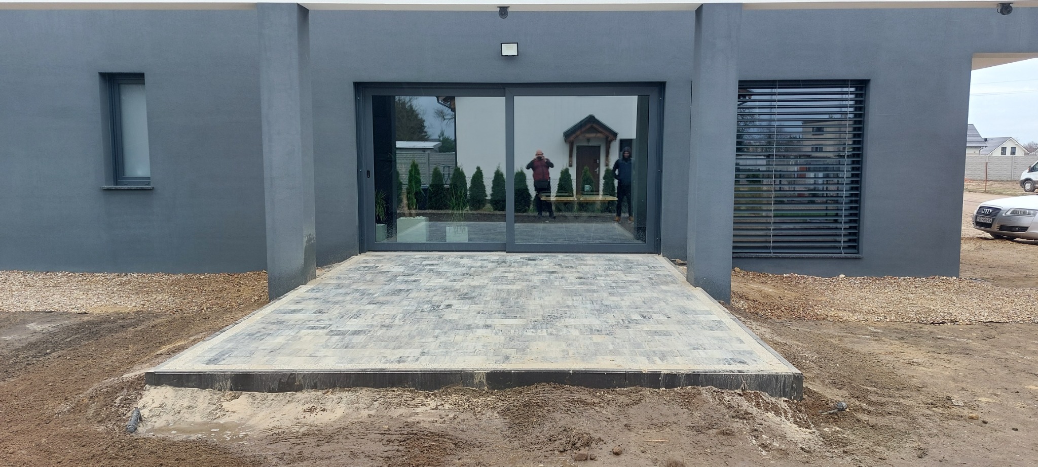 Szary, nowoczesny budynek z nowo ułożoną kostką brukową na podejściu do szklanych drzwi przesuwnych, widoczne odbicia osób w szybach.