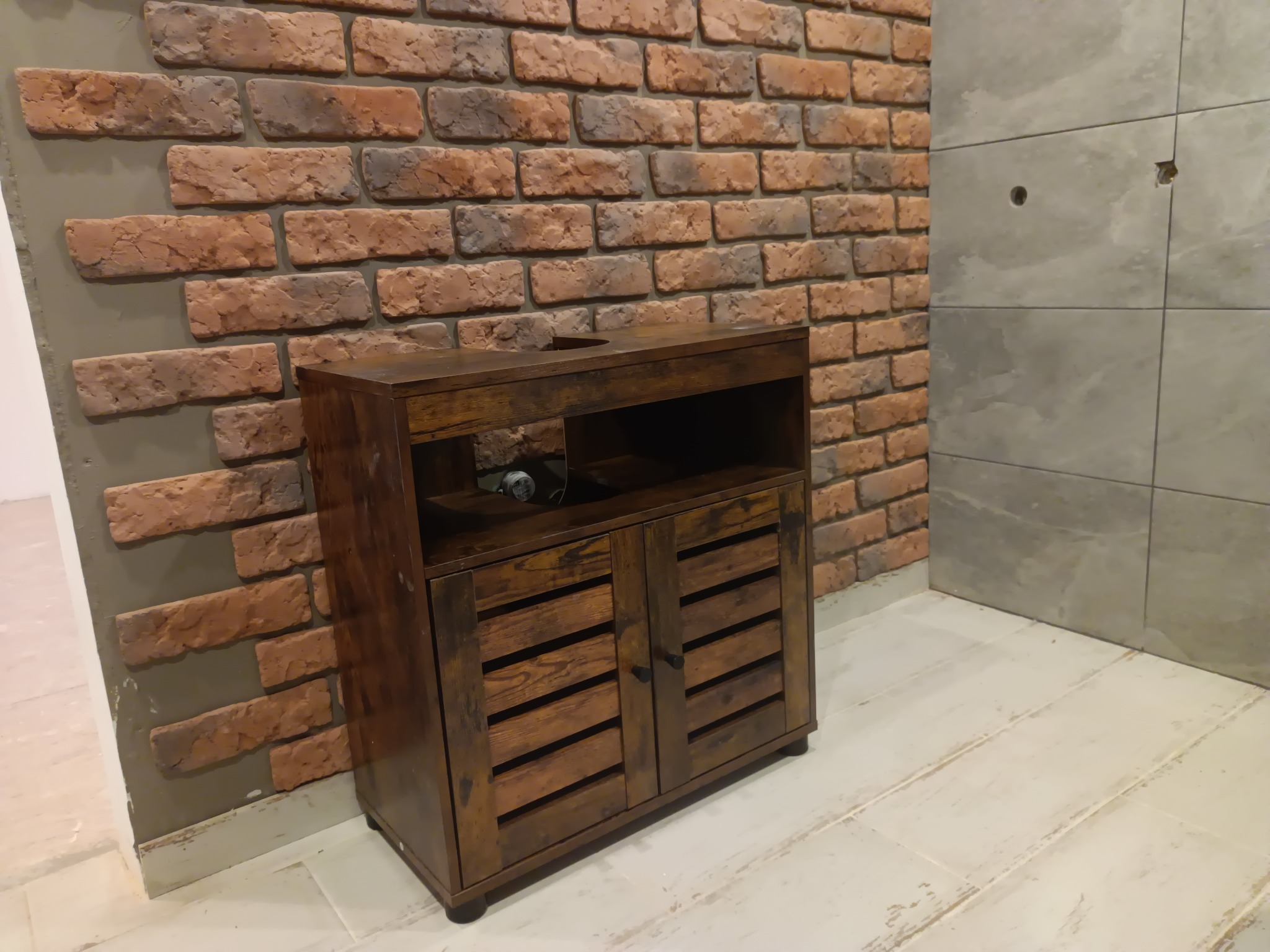 Ciemnobrązowa szafka pod umywalkę z lamelowymi drzwiczkami, na tle ściany z cegły klinkierowej i szarych płytek, widoczne otwory po montażu.