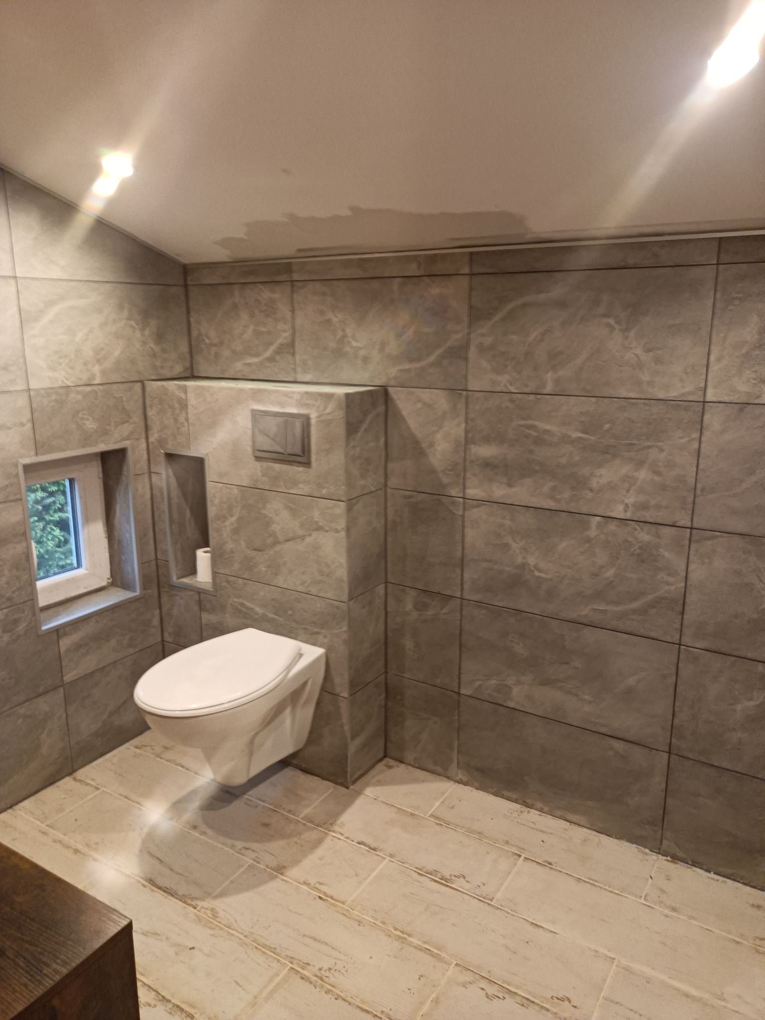 Nowoczesna łazienka z szarymi płytkami imitującymi kamień, podwieszana toaleta, małe okno i wnęka na papier toaletowy.