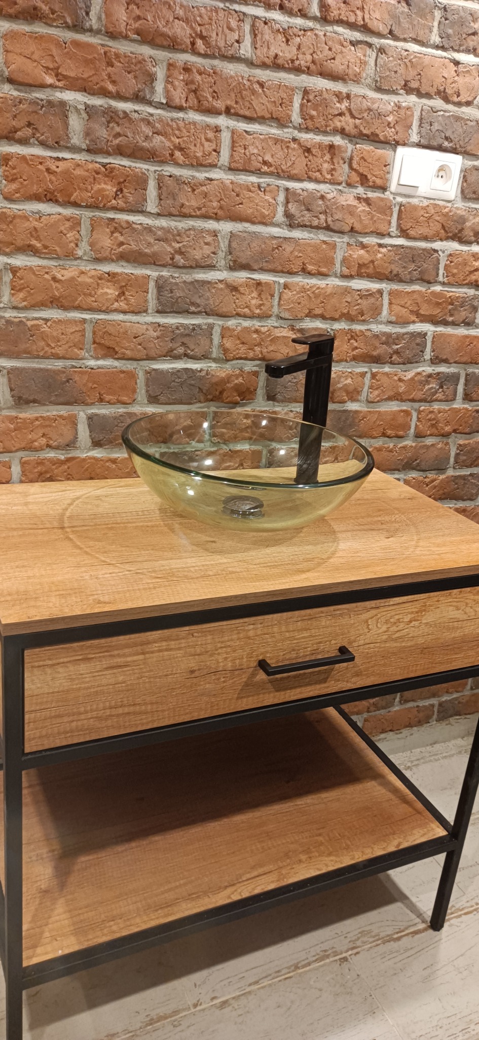 Szklana umywalka nablatowa z czarną armaturą na drewnianej szafce z szufladą, tło z cegły klinkierowej z gniazdkiem elektrycznym.