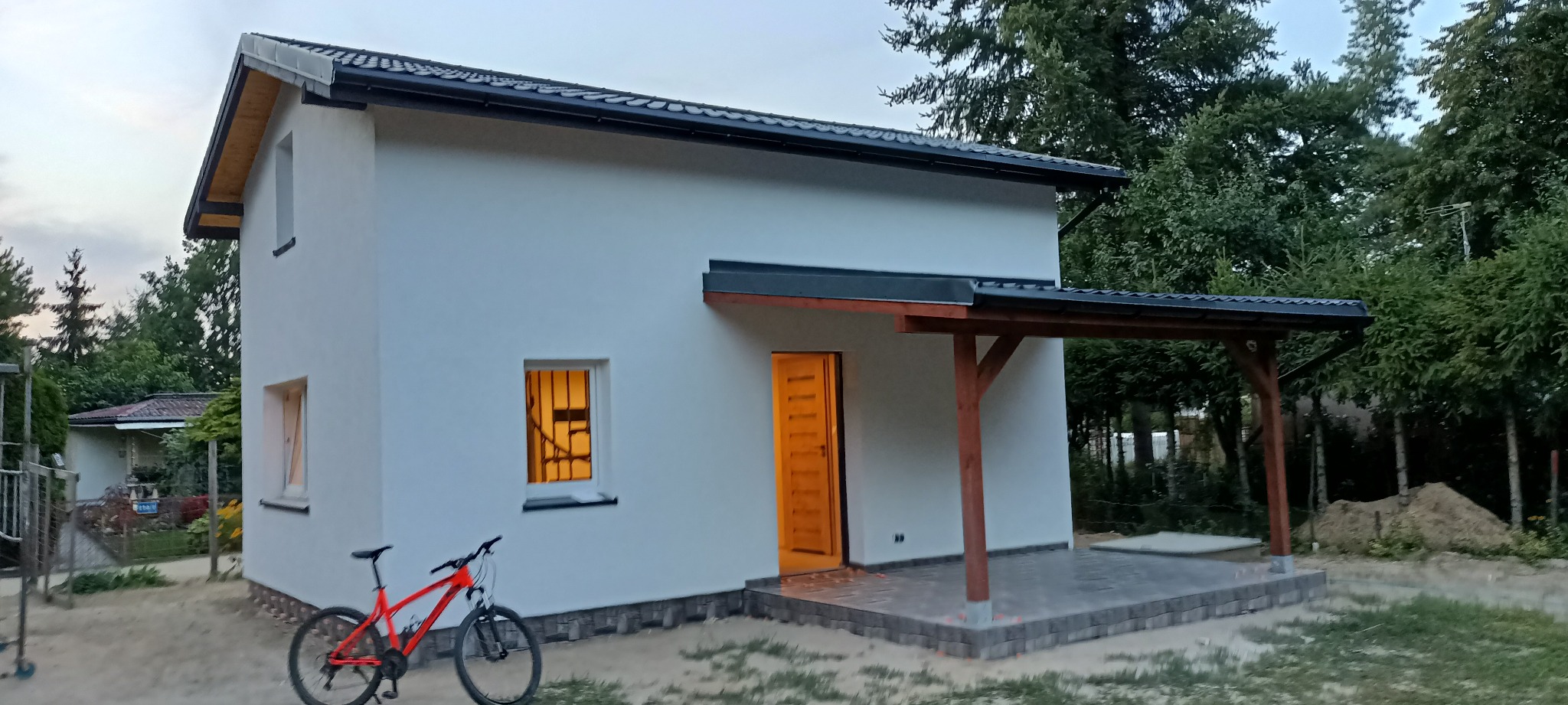 Nowy, parterowy dom z białymi ścianami, czarnym dachem i drewnianą wiatą, z rowerem górskim w kolorze czerwonym zaparkowanym obok, widok na otwarte drzwi i oświetlone wnętrze.