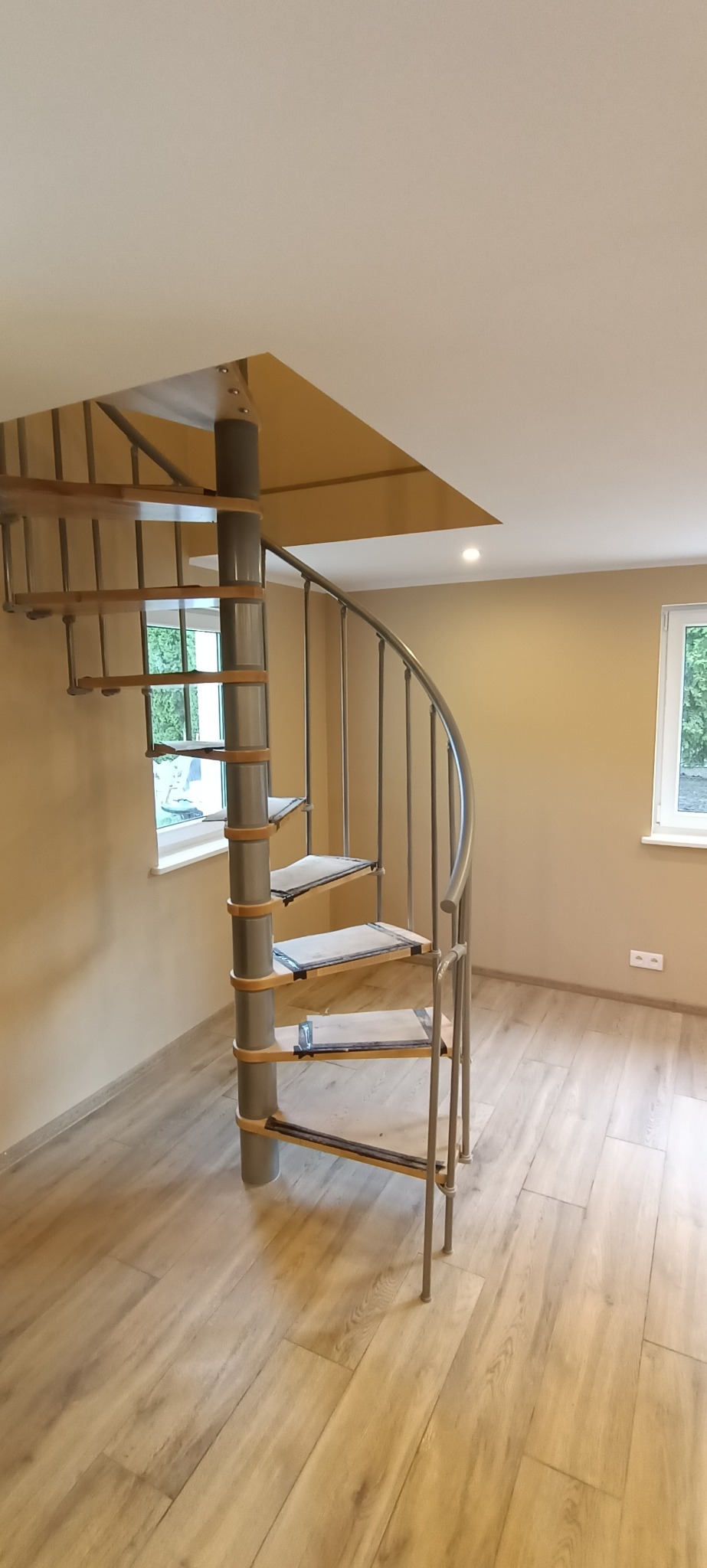 Metalowe schody spiralne z drewnianymi stopniami w jasnym wnętrzu z panelami podłogowymi i beżowymi ścianami.
