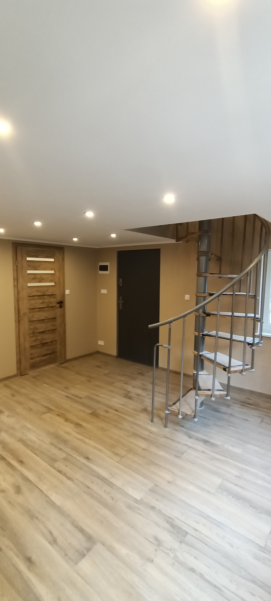 Jasne wnętrze po renowacji z drewnopodobną podłogą, spiralnymi schodami, dwojgiem drzwi i oświetleniem punktowym w suficie.