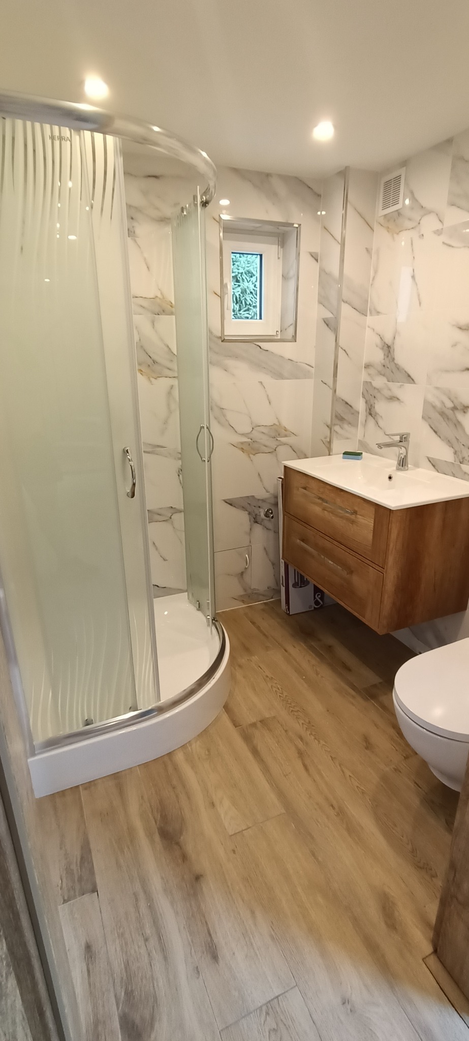 Nowoczesna łazienka z kabiną prysznicową z ornamentem na szkle, drewnianą szafką pod umywalką i marmurowymi płytkami na ścianach, widok z perspektywy podłogi