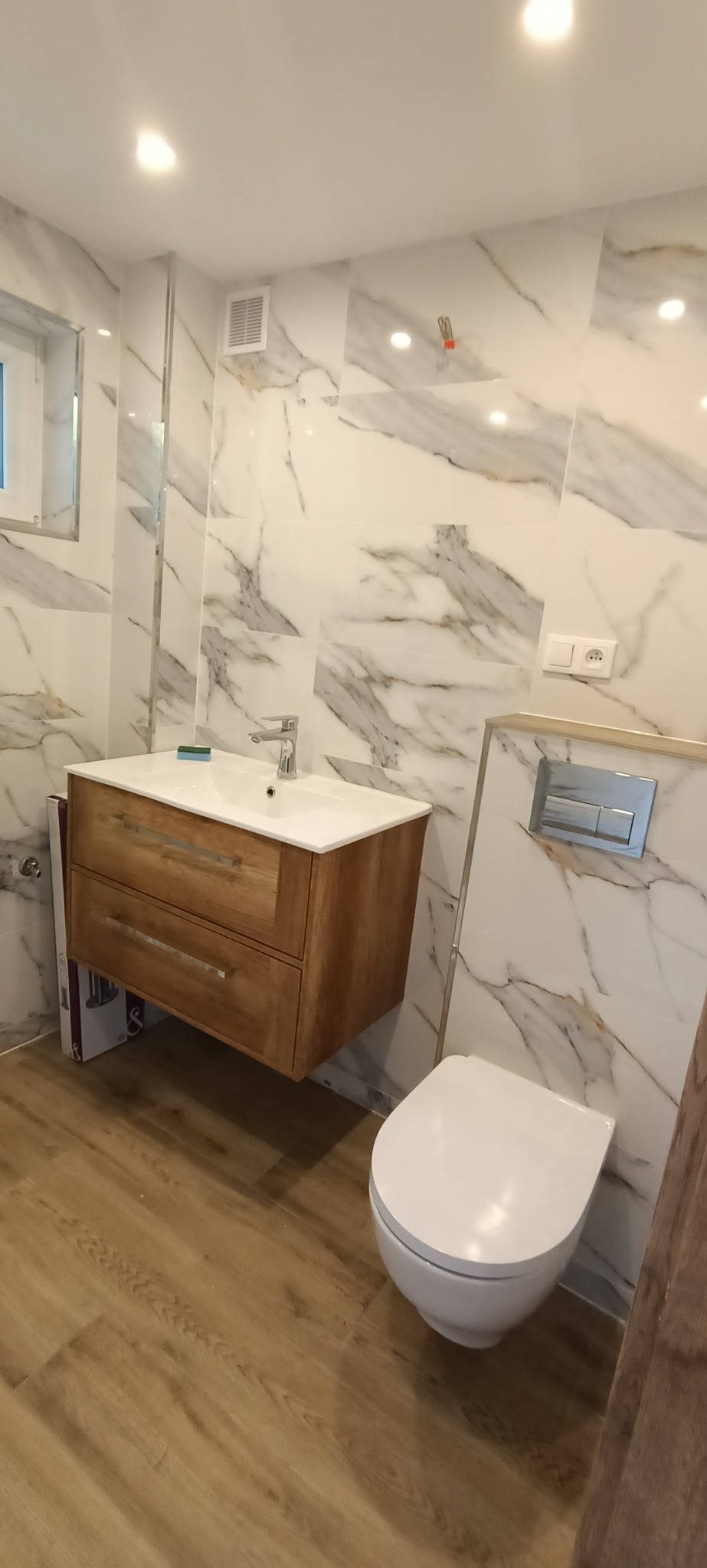 Nowoczesna łazienka z marmurowymi płytkami, drewnianą szafką pod umywalką i podwieszaną toaletą z chromowanym przyciskiem spłukiwania, oświetlona punktowymi lampami sufitowymi.