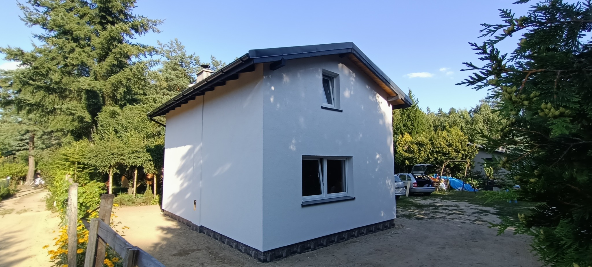 Otynkowany, biały budynek gospodarczy z dwoma oknami, czarnym dachem i wykończeniem z imitacji kamienia u podstawy, otoczony zielenią i z samochodem w tle.