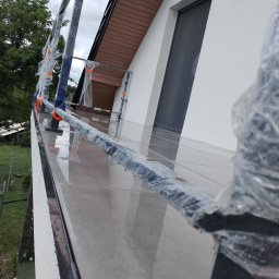 Max Construction - Montaż obróbek blacharskich na nowym budynku, widoczne rusztowanie zabezpieczone folią, dach z brązową podbitką i fragment elewacji z oknem.