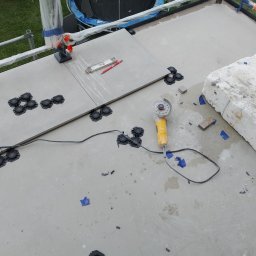 Max Construction - Montaż płyt tarasowych na regulowanych wspornikach, widoczna poziomica, ołówek, szlifierka kątowa i fragment rusztowania zabezpieczonego folią.