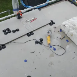 Montaż płyt tarasowych na regulowanych wspornikach, widoczna poziomica, ołówek, szlifierka kątowa i fragment rusztowania zabezpieczonego folią.