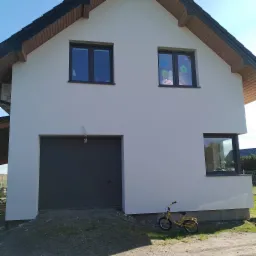 Dwukondygnacyjny dom jednorodzinny z szarą bramą garażową i żółtym rowerem dziecięcym opartym o ścianę, okna z ciemnymi ramami, elewacja w jasnym odcieniu, dach pokryty czarną dachówką.