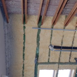 Max Construction - Poddasze ocieplone pianą PUR, widoczne drewniane krokwie, zielone elementy konstrukcyjne, instalacja wentylacyjna z czarnymi rurami karbowanymi oraz okno dachowe w dolnej części kadru.