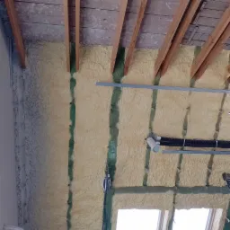 Poddasze ocieplone pianą PUR, widoczne drewniane krokwie, zielone elementy konstrukcyjne, instalacja wentylacyjna z czarnymi rurami karbowanymi oraz okno dachowe w dolnej części kadru.