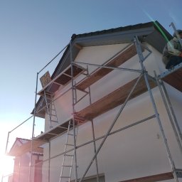 Max Construction - Dom jednorodzinny w trakcie prac elewacyjnych, widoczne rusztowanie, częściowo wykończona elewacja i pracownik w górnej części rusztowania, na dachu szara blachodachówka.