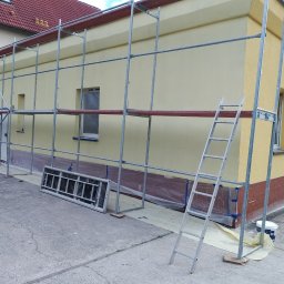 Max Construction - Żółta elewacja budynku w trakcie malowania, widoczne rusztowanie, zabezpieczone okna folią, metalowa drabina oparta o ścianę, wiadro z farbą u podstawy rusztowania.