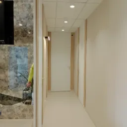 Korytarz biurowy z białymi ścianami, sufitem podwieszanym z oświetleniem punktowym i ukrytymi drzwiami, wykończony elementami drewnopodobnymi.