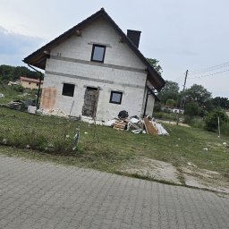 Max Construction - Surowy dom z białych bloczków, czarną stolarką i dachem, w trakcie budowy. Widoczne materiały budowlane i teren wokół. Ujęcie z poziomu chodnika.