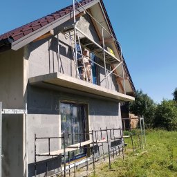 Max Construction - Dom w trakcie ocieplania styropianem, widoczne rusztowania i pracownik na wysokości. Dach z brązowej dachówki, okna zabezpieczone folią.