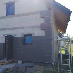 Max Construction - Fragment ocieplanej ściany domu z szarego styropianu, widoczne okna zabezpieczone folią, drabina i porozrzucane płyty izolacyjne. Częściowe ocieplenie elewacji.
