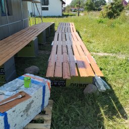 Max Construction - Malowanie desek elewacyjnych na zewnątrz domu. Deski ułożone na rusztowaniu, obok puszka farby i pędzle. W tle dom z ocieploną elewacją i rusztowaniem.