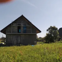 Surowy, dwukondygnacyjny dom w trakcie budowy, z szarymi ścianami i rusztowaniami; wysoka trawa na pierwszym planie, błękitne niebo w tle. Widok z poziomu gruntu.