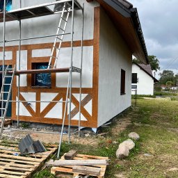 Max Construction - Dom w trakcie prac elewacyjnych: widoczne rusztowanie, fragmenty drewnianej konstrukcji szkieletowej i otynkowana ściana. Palety leżą na trawie.